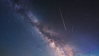 Milky way looks like a large cloudy soup of stars stretching across the sky. There are a few streaks of white lights across the scene, these are meteors.