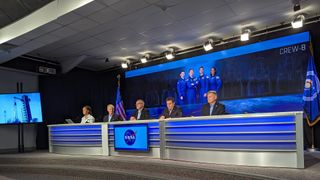 four people in black suits with blue shirts sit at a table in front of a poster of four astronauts in blue flight suits and the text "Crew-8"